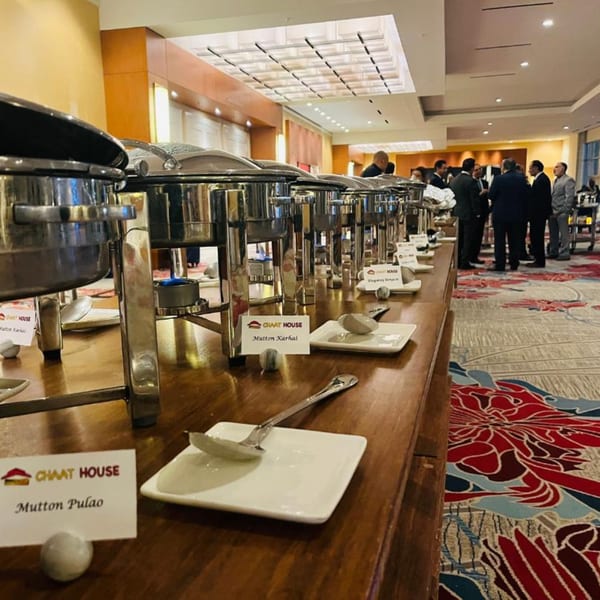 Chaat House buffet line with chafing dishes, serving utensils, and men in suits in the background.