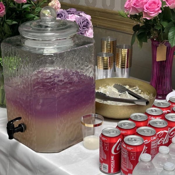 A beverage dispenser with purple drink, ice bucket, Coca-Cola cans, and pink roses on a table.