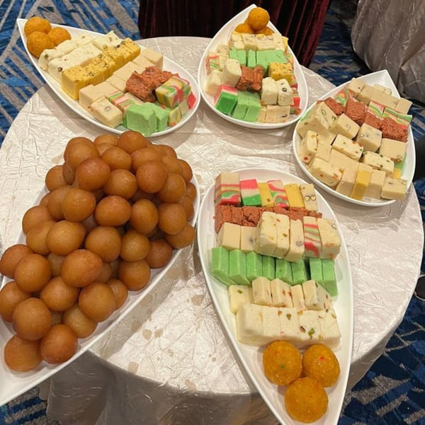 A table laden with white platters of colorful Pakistani sweets, including gulab jamun and barfi.