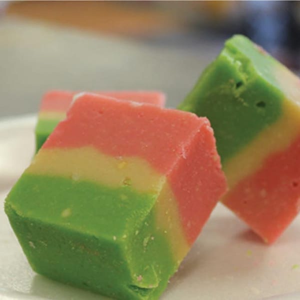 Three colorful Pakistani barfi sweets, layered in green, yellow, and pink, on a white surface.