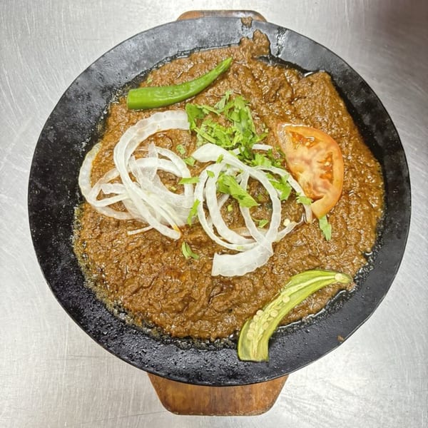 Pakistani dish with brown curry, sliced onions, cilantro, tomato, and green chilies in a black pan.